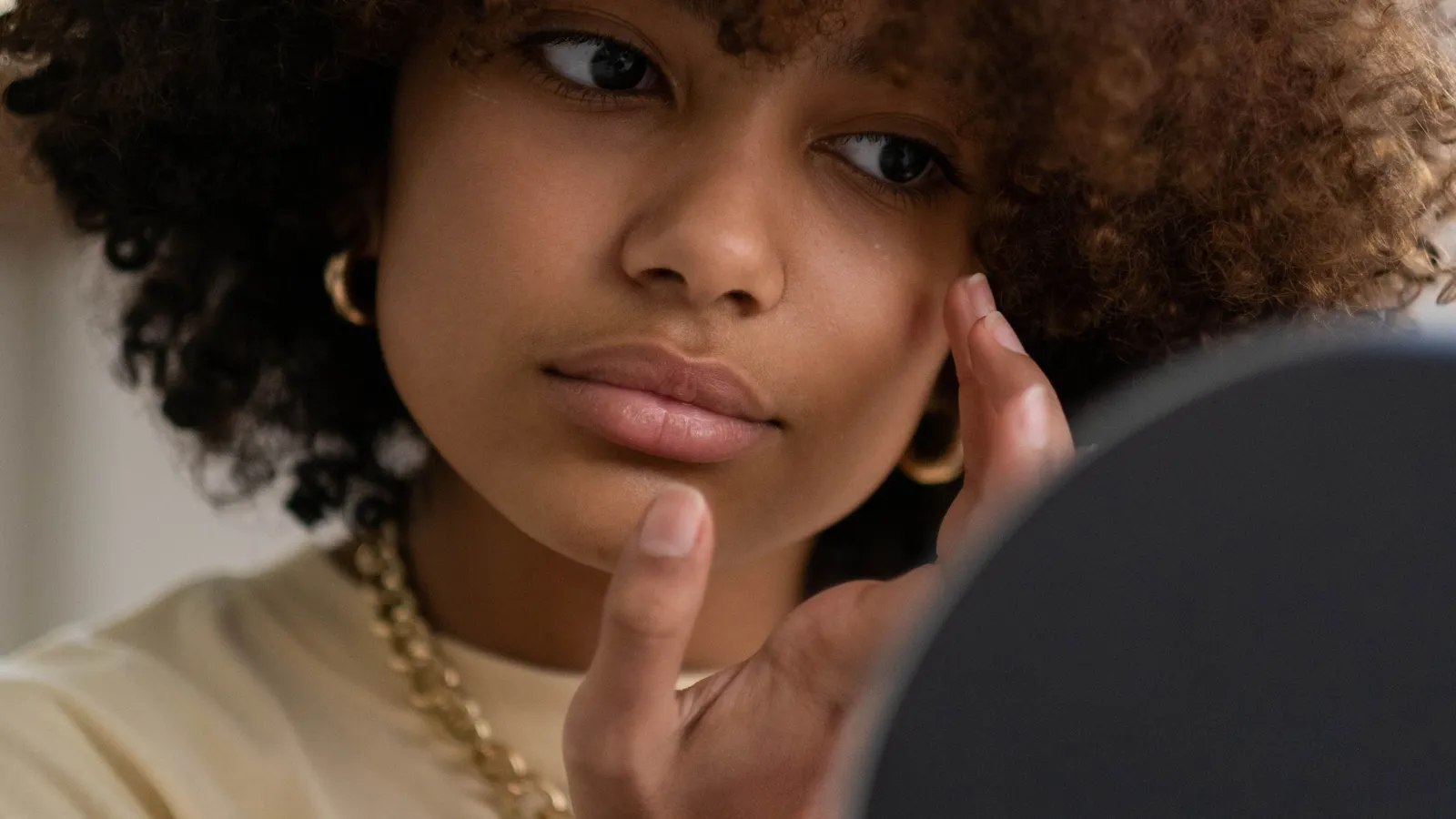 A woman with natural hair checks her skin in a handheld mirror.