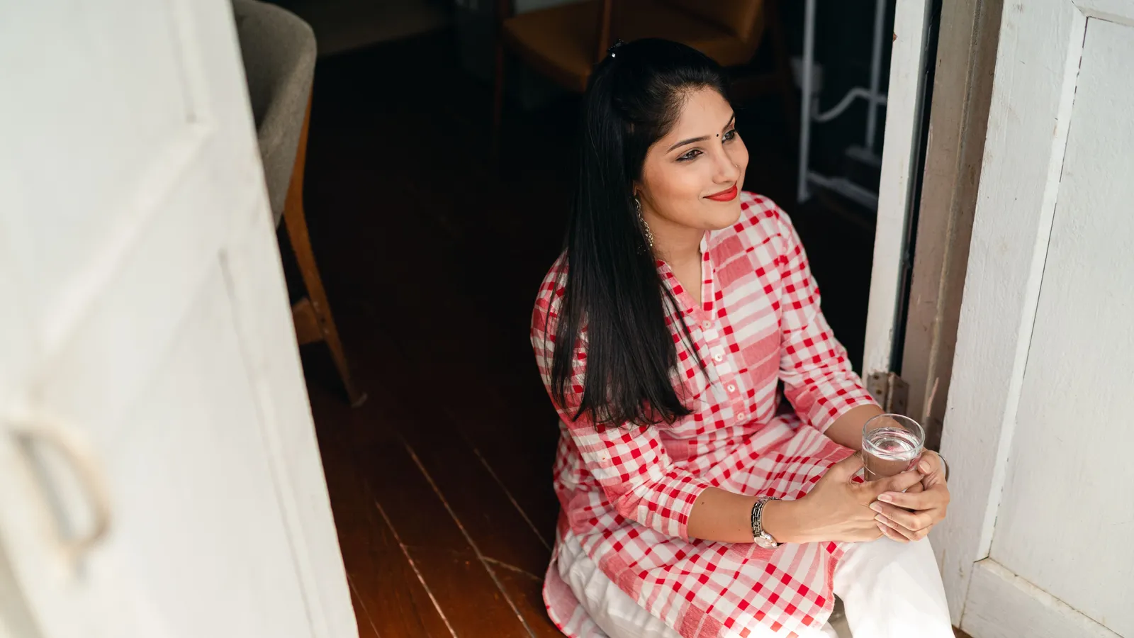 A woman sits in a sunny doorway with a glass of water.